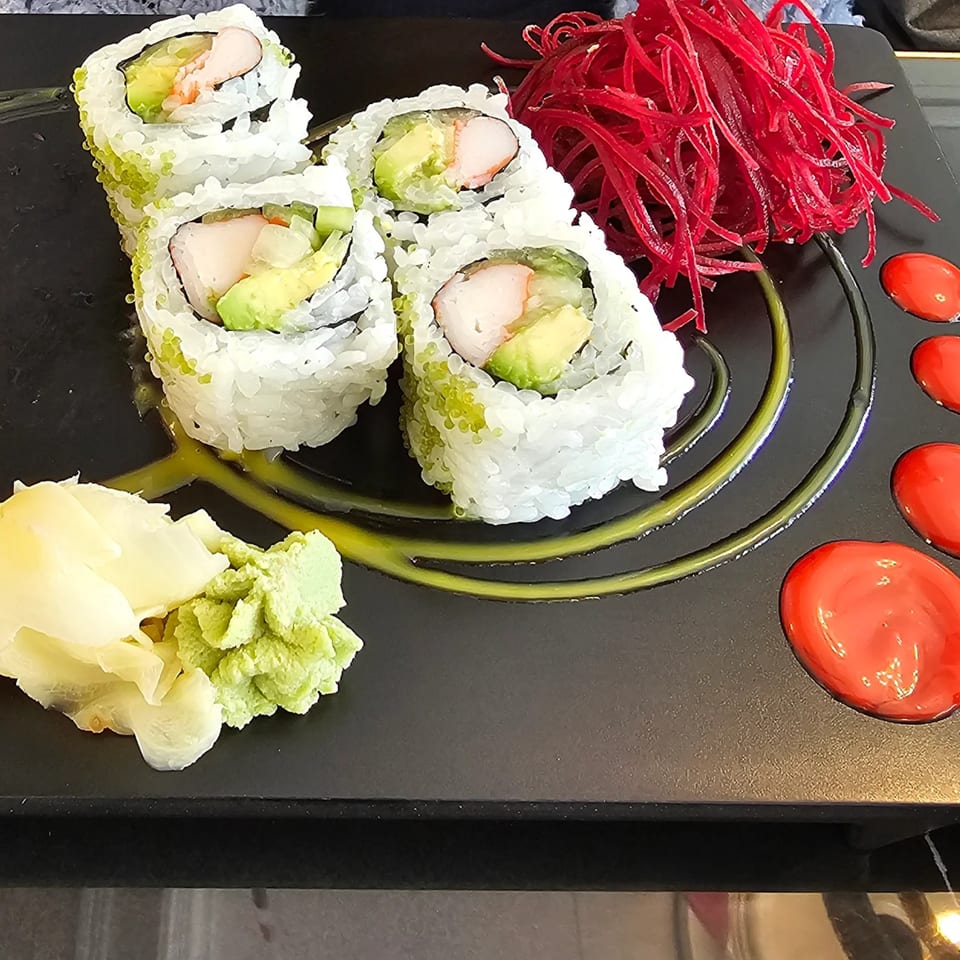 Four California rolls with green tobiko, red beet strings, wasabi, and ginger on a black plate.