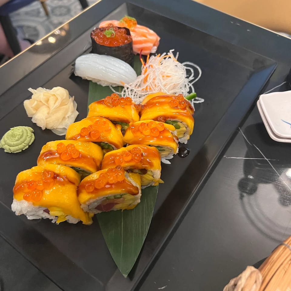 A black platter of sushi rolls topped with mango and roe, with sashimi and garnishes.