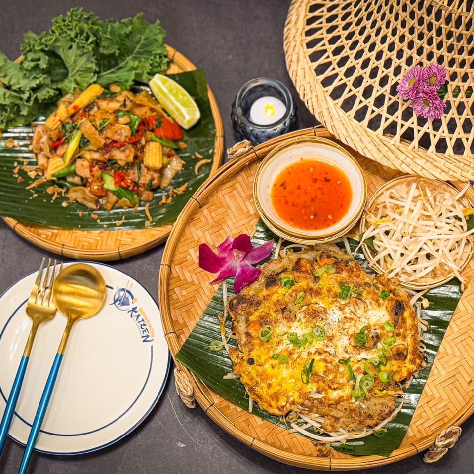 Two Thai dishes, one with noodles and vegetables, the other an omelet, on woven platters.