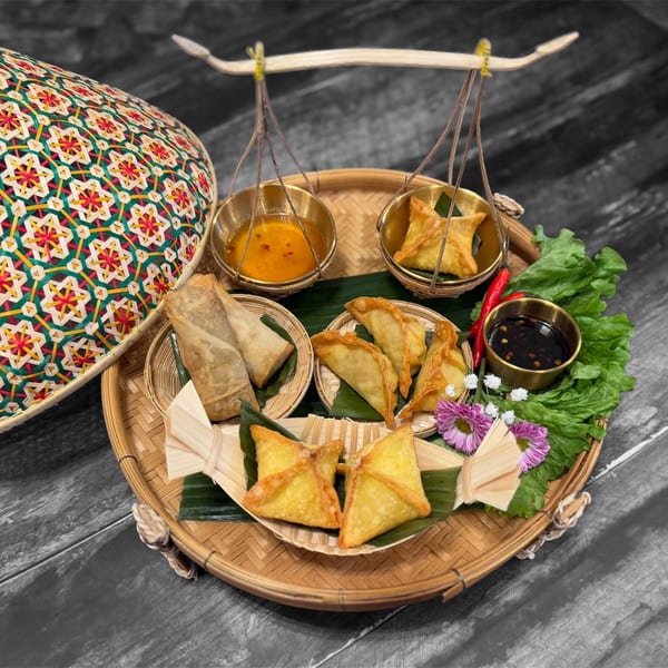 A bamboo tray with various Thai appetizers, including spring rolls, dumplings, and crab rangoons, with dipping sauces.