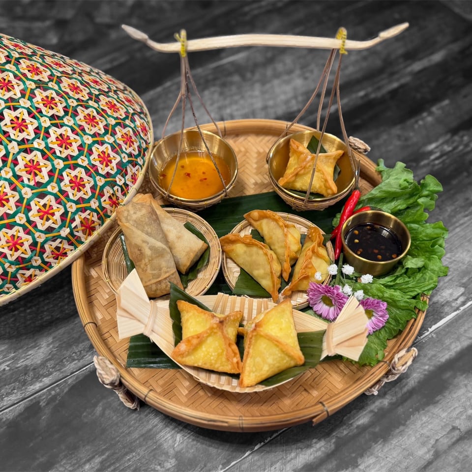 A bamboo tray with various Thai appetizers, including spring rolls, dumplings, and crab rangoons, with dipping sauces.