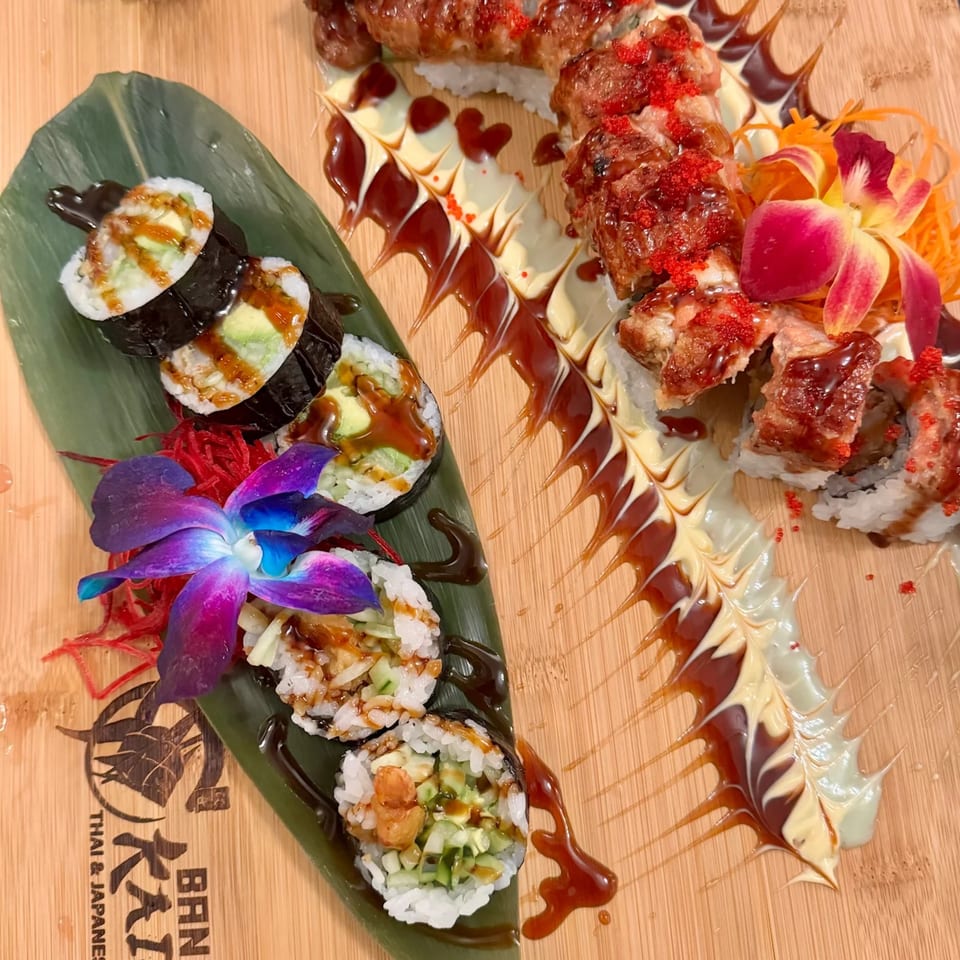 Sushi rolls with avocado, shrimp, and eel, garnished with orchids on a wooden board.