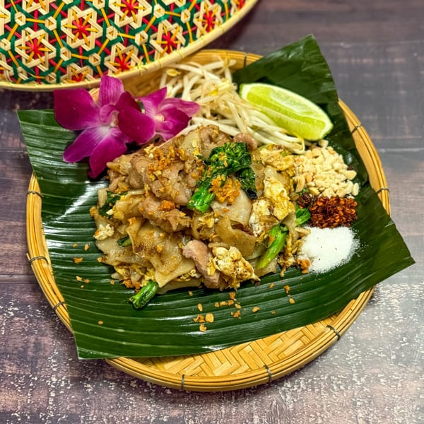 Pad See Ew with pork, egg, and greens, garnished with lime, sprouts, and spices on a banana leaf.
