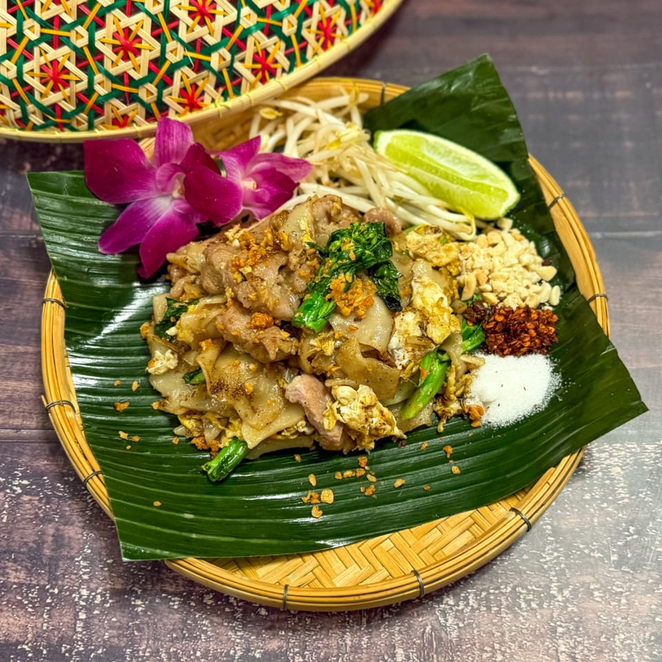 Pad See Ew with pork, egg, and greens, garnished with lime, sprouts, and spices on a banana leaf.