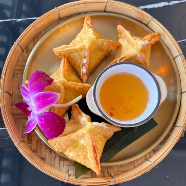 Golden fried wontons with a sweet chili dipping sauce and a purple orchid on a bamboo tray.