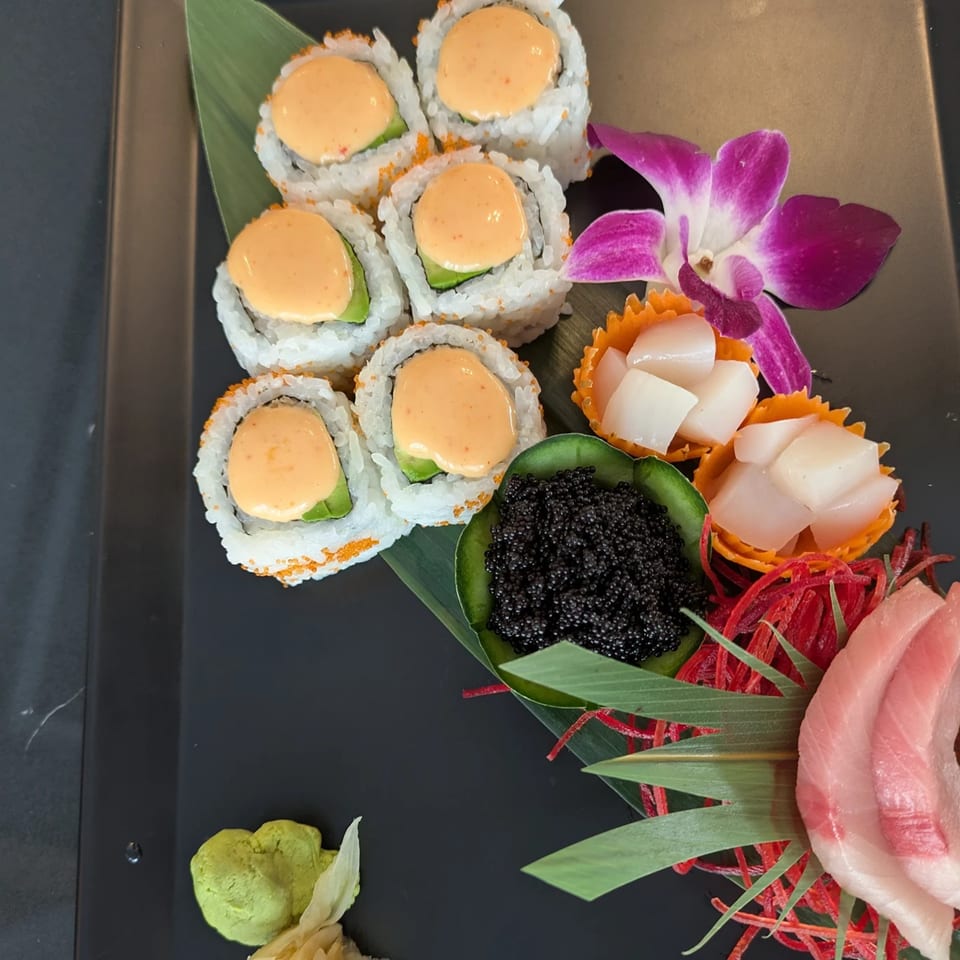 Sushi rolls with orange sauce, caviar, scallops, tuna sashimi, wasabi, and ginger on a black plate.