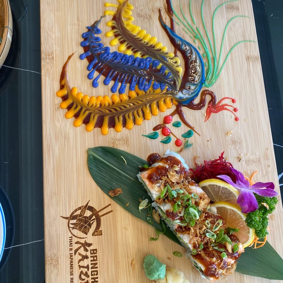 Sushi rolls on a bamboo board with a colorful sauce design and the Bangkok Kaizen logo.