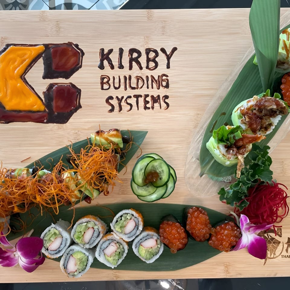 Sushi and rolls on a wooden board, with "Kirby Building Systems" written in sauce.